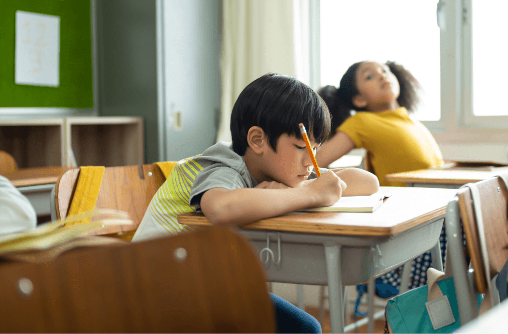 Young boy focusing on his writing in a classroom with a classmate in the background.